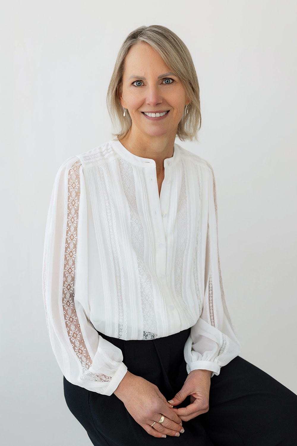 Smiling woman with short blond hair wearing a white lace blouse and black pants, seated against a light backdrop.