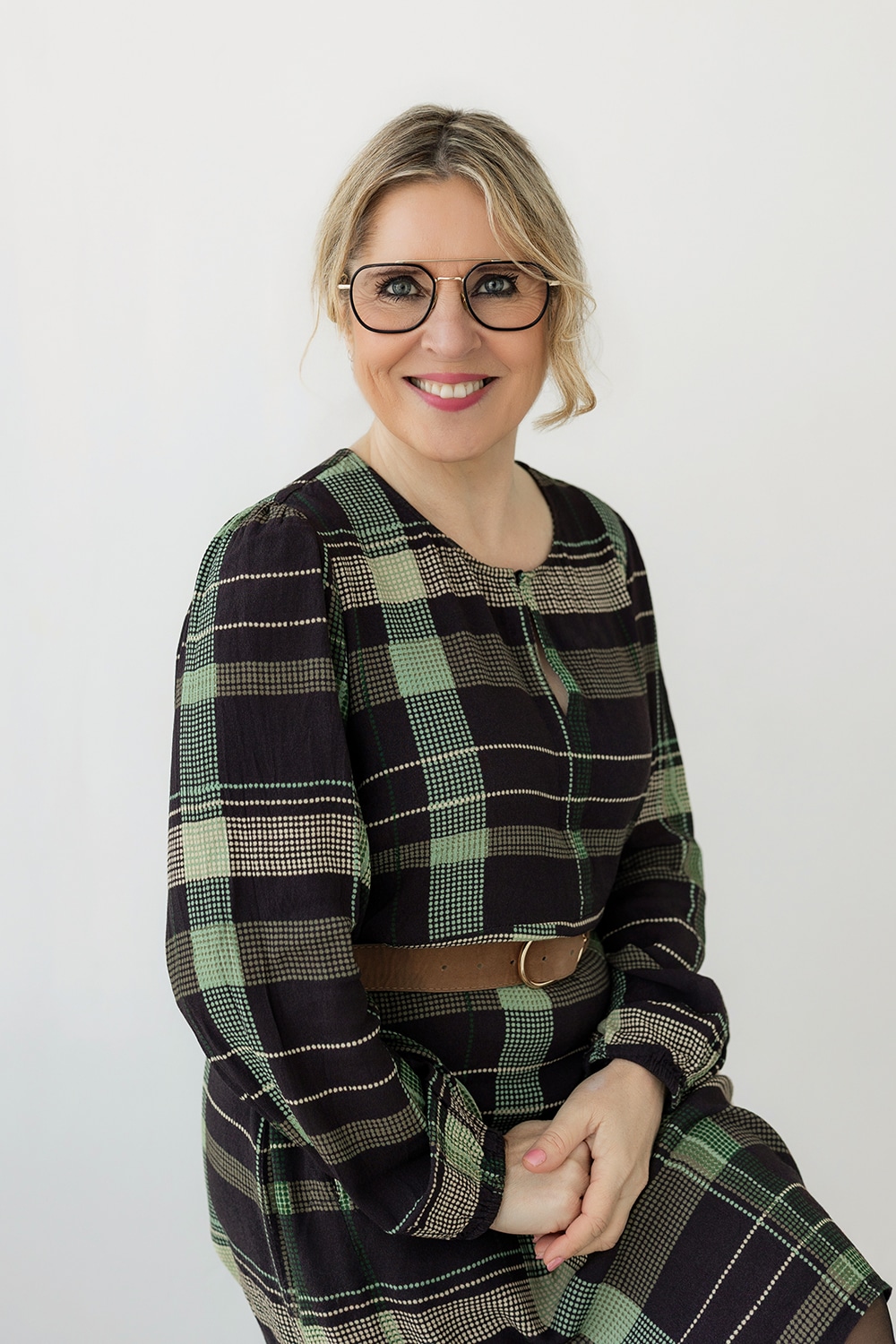 Smiling woman with short blond hair and round glasses, wearing a green plaid dress with a brown belt, against a light background.